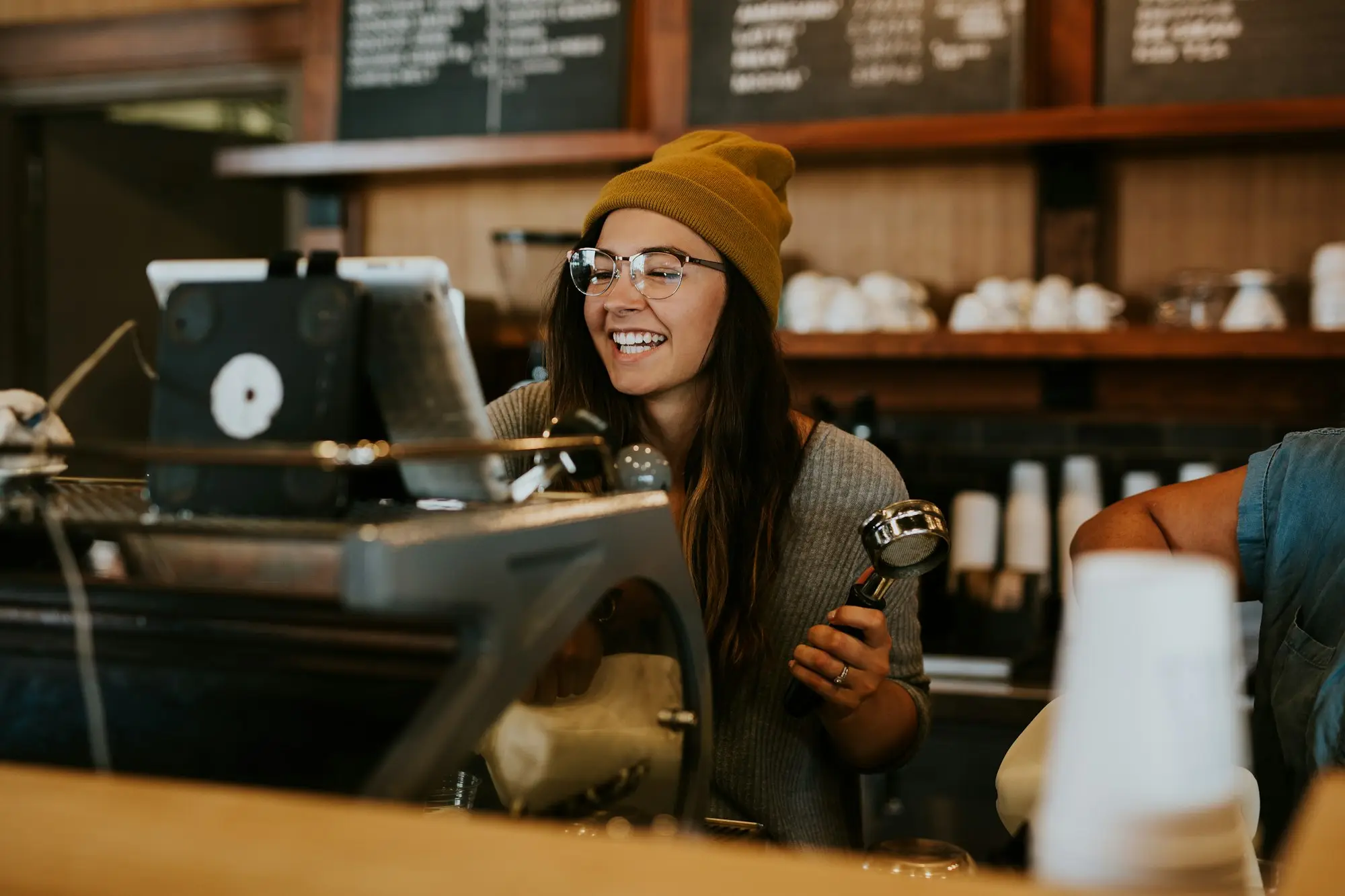 Smiling barista in a mustard beanie holding a portafilter beside the espresso machine