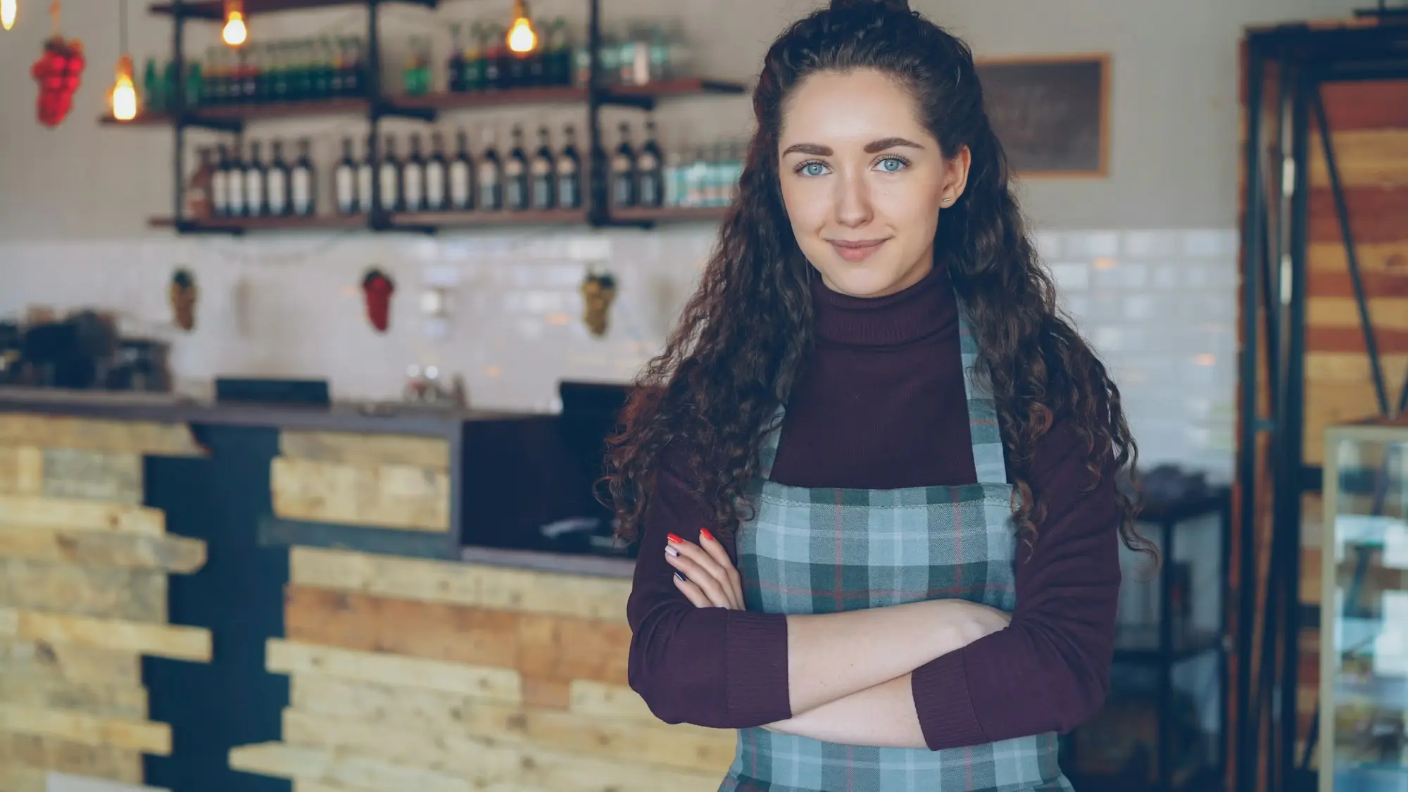Cafe owner in a checked apron standing with arms folded in front of her coffee counter