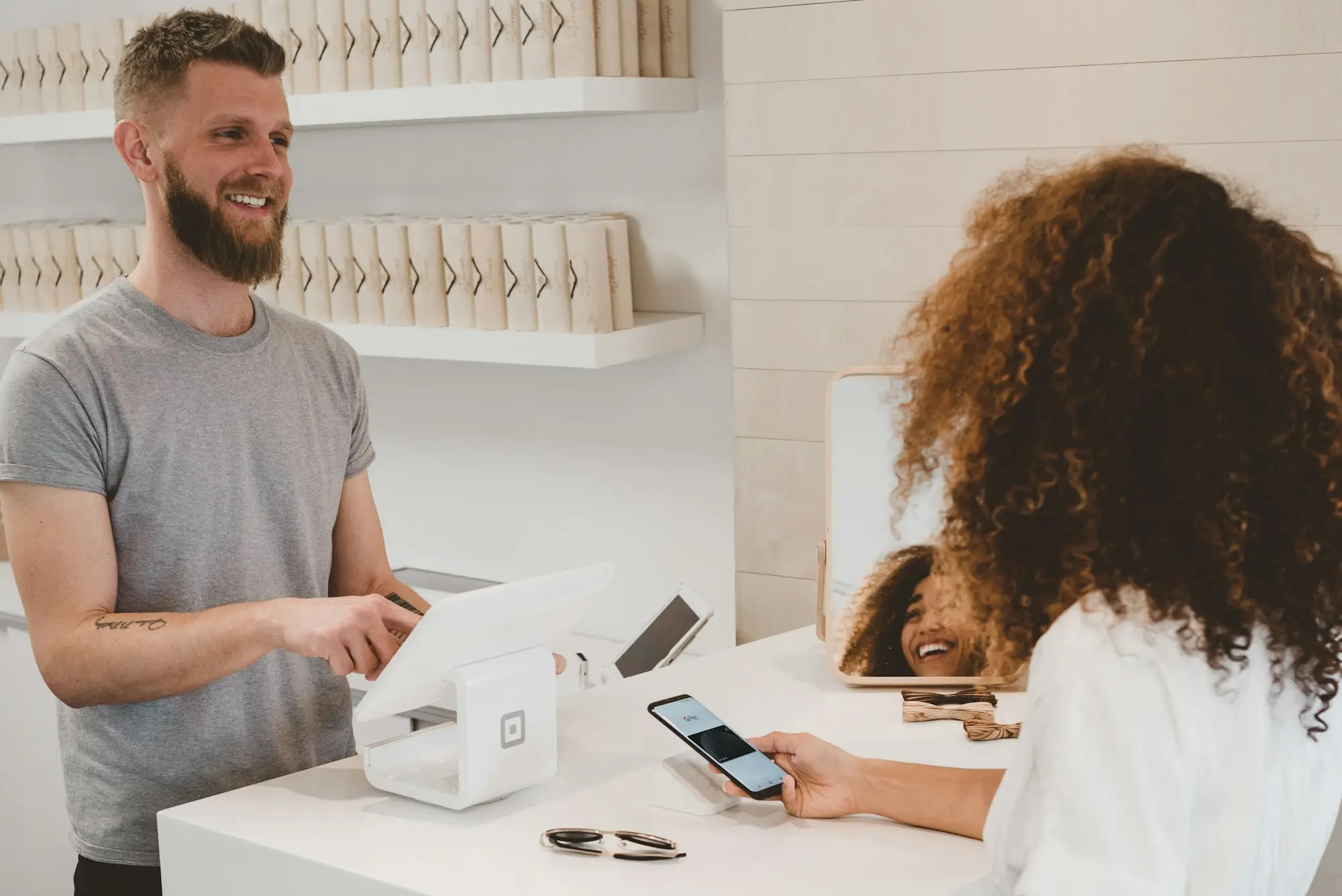 Customer tapping a phone to pay at a small shop counter while the owner rings up the sale