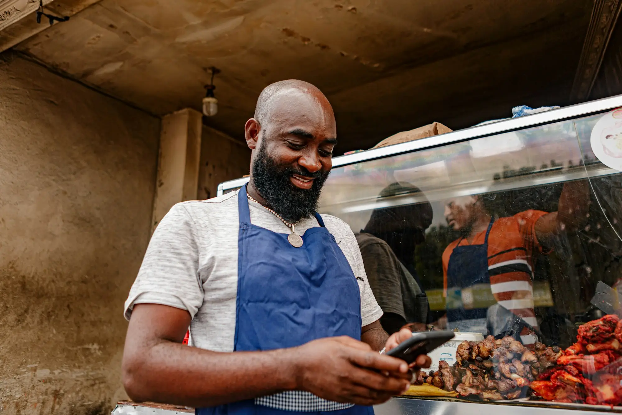 Deli owner in a blue apron smiling at his phone behind a glass food display counter