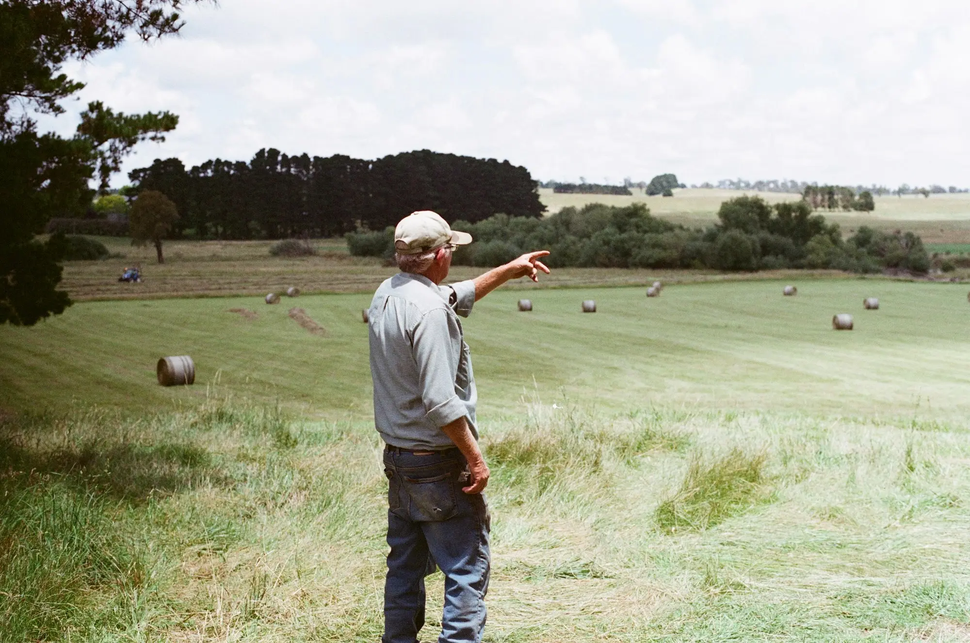 Farmer in a cap pointing across a paddock dotted with round hay bales