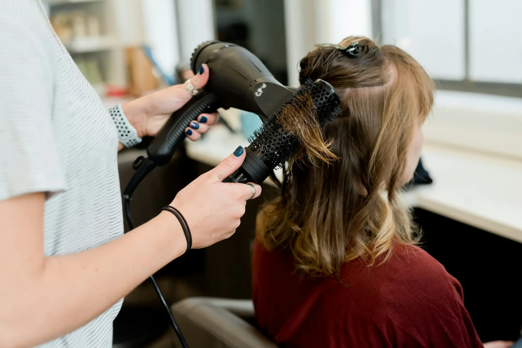 Hairstylist blow-drying a client's hair with a round brush in a sunny salon