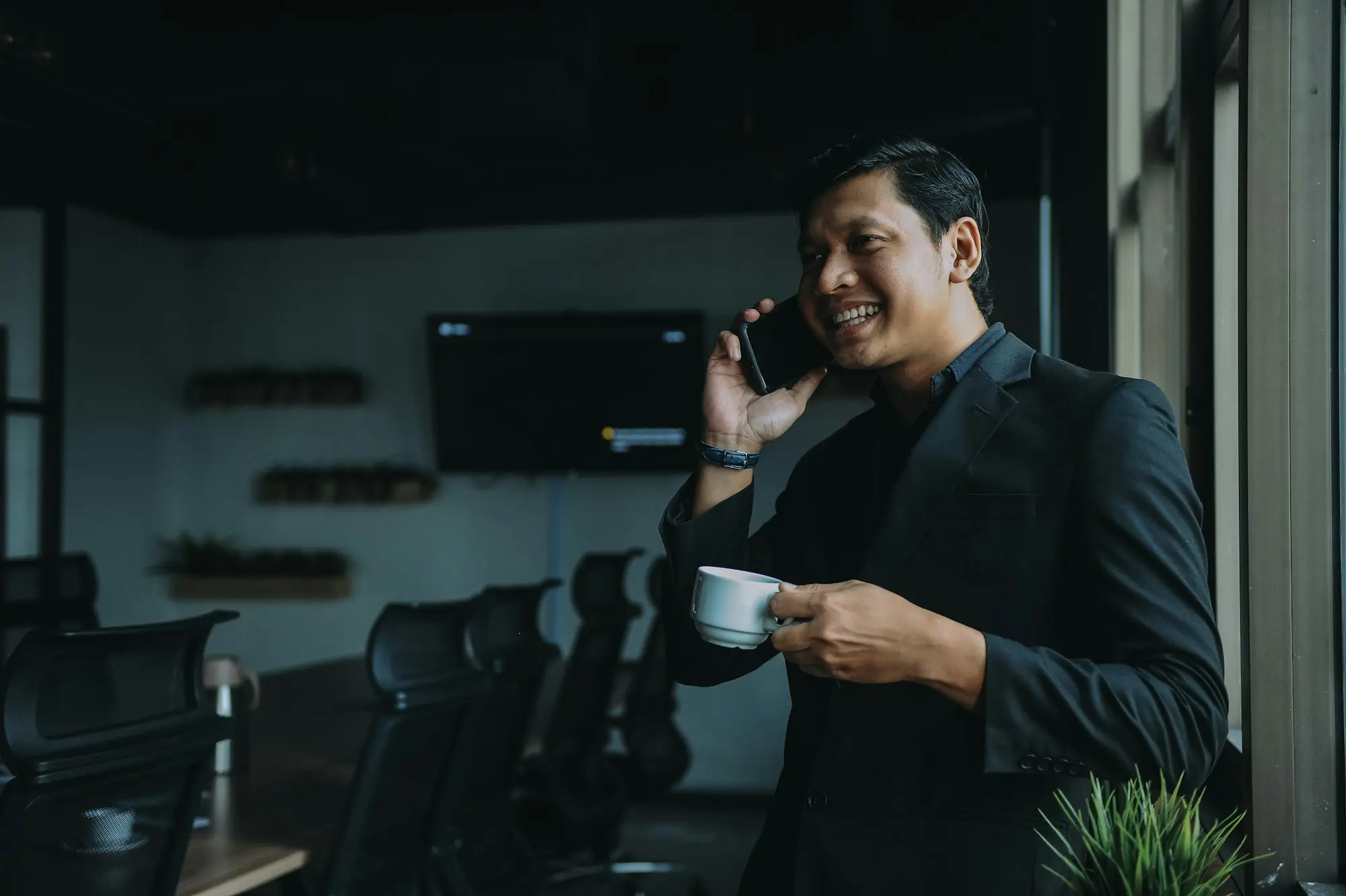 Business manager smiling on a phone call with a coffee cup in a dark modern office
