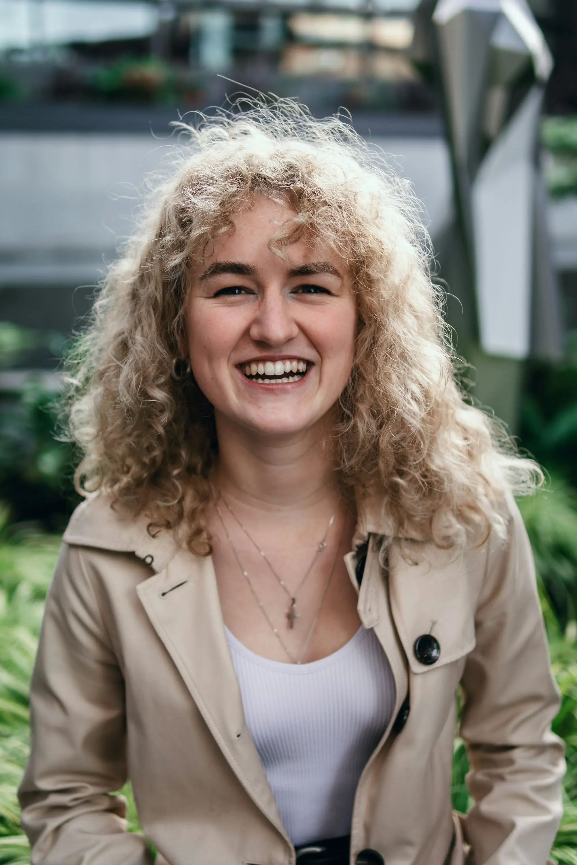 Business owner with curly blonde hair in a beige coat laughing outdoors among green plants