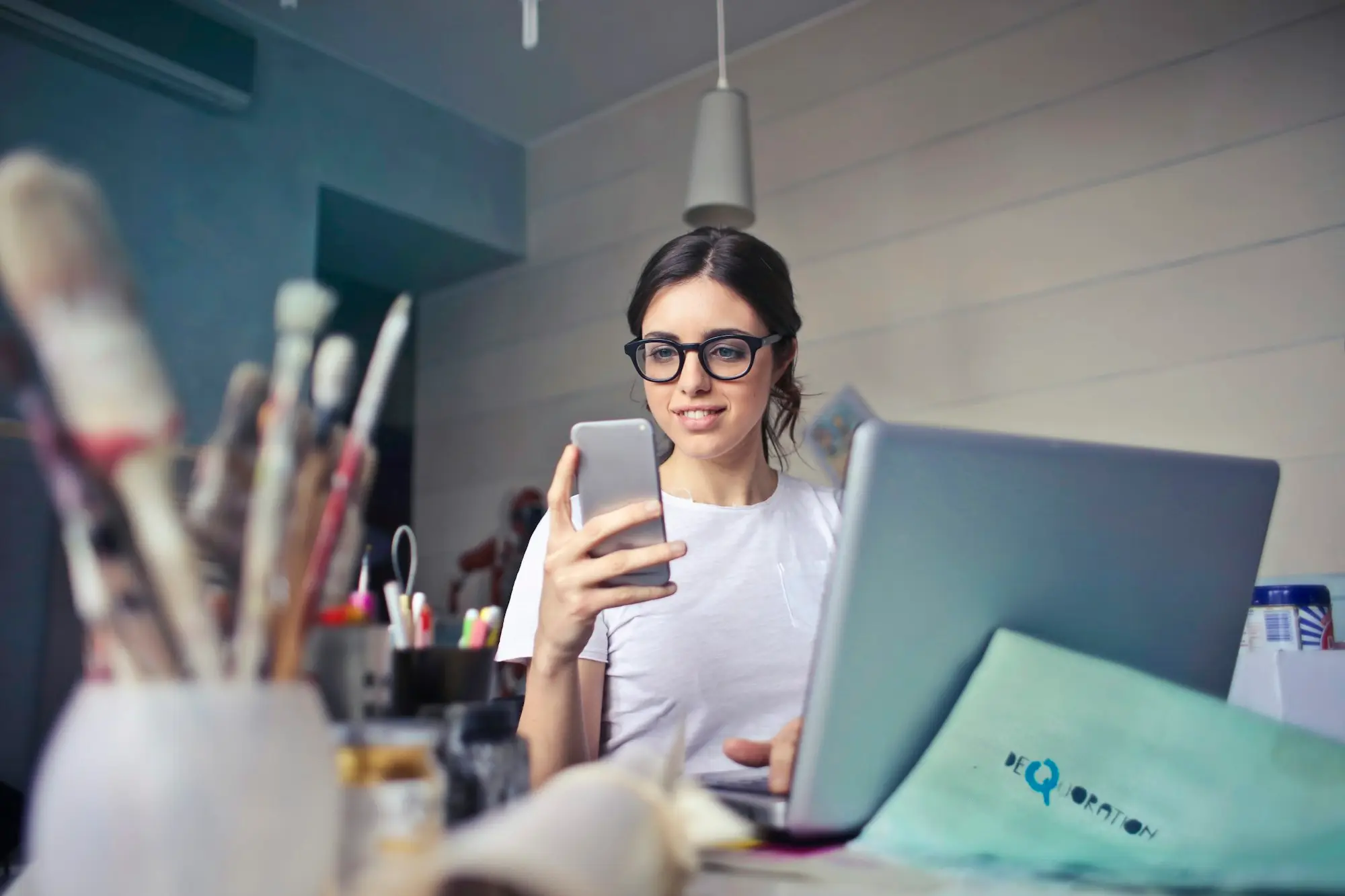 Small business owner in glasses checking her phone beside a laptop in a bright studio workspace