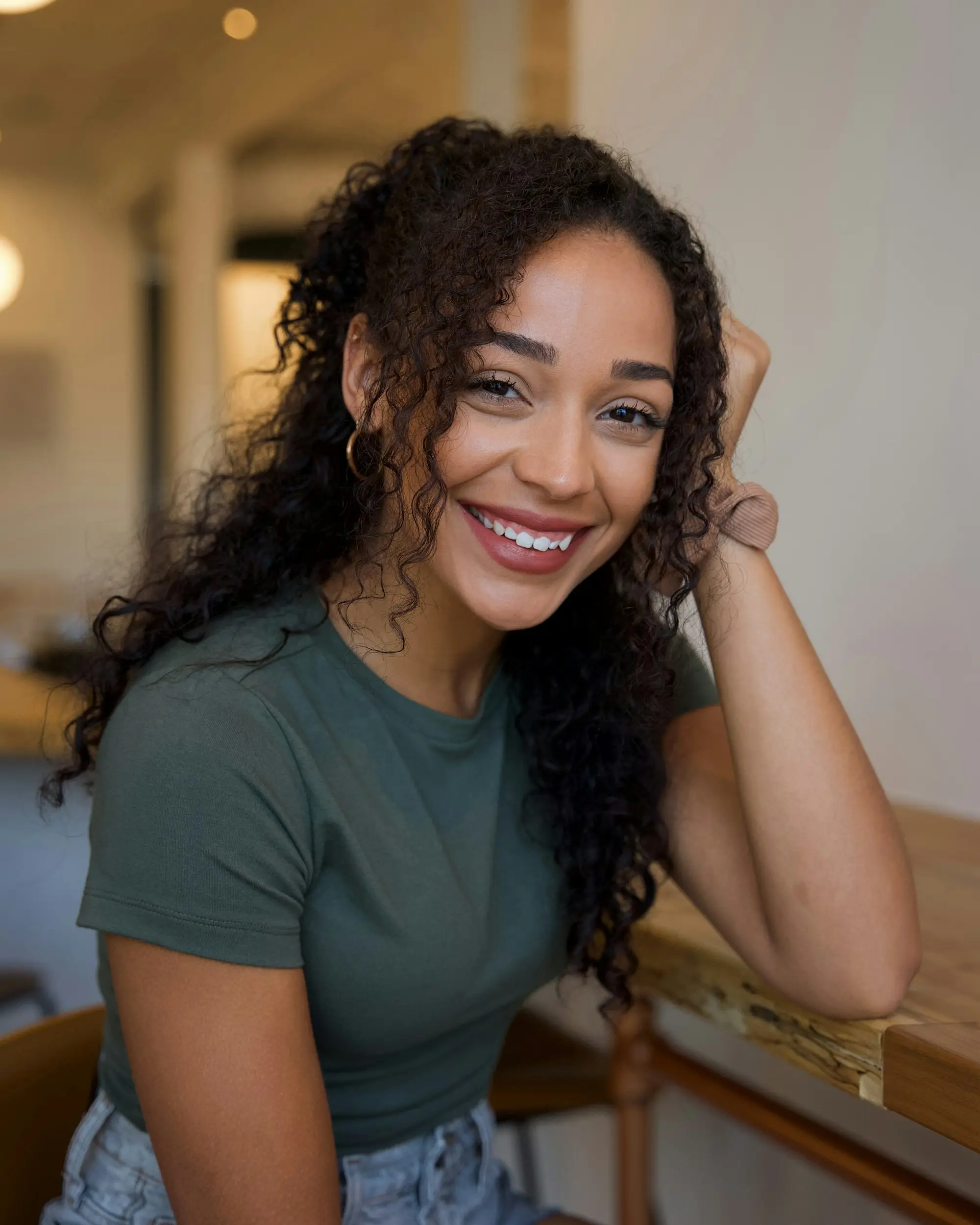 Smiling business owner with curly hair in a green t-shirt leaning on a timber counter in her cafe