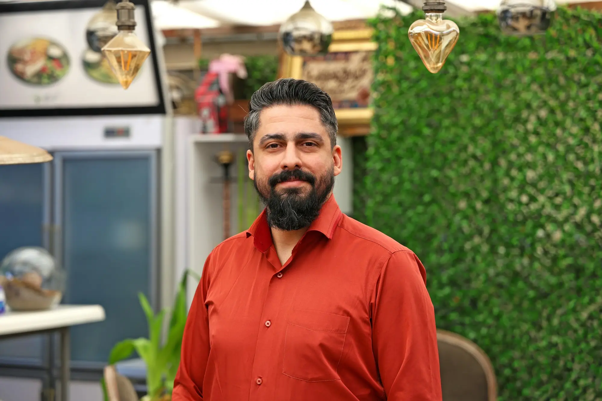 Restaurant owner with a beard in a red shirt smiling in his dining room beside a green wall