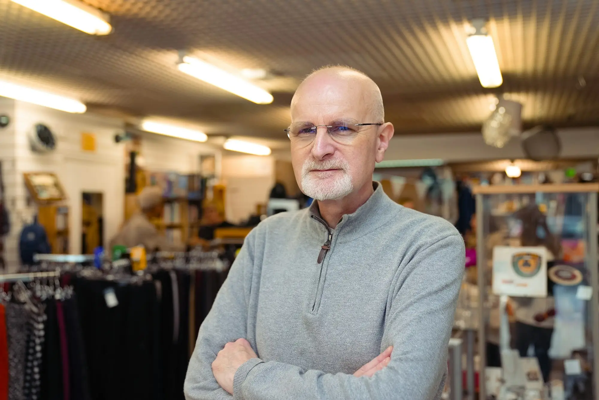 Retail store owner in a grey jumper standing with arms folded among racks of stock