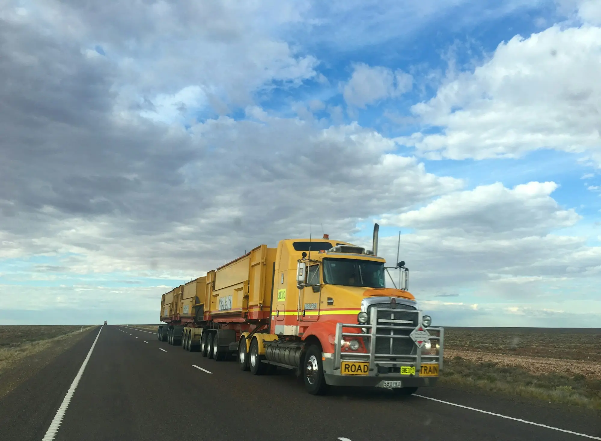 Road train hauling freight along a long outback highway under a wide cloudy sky