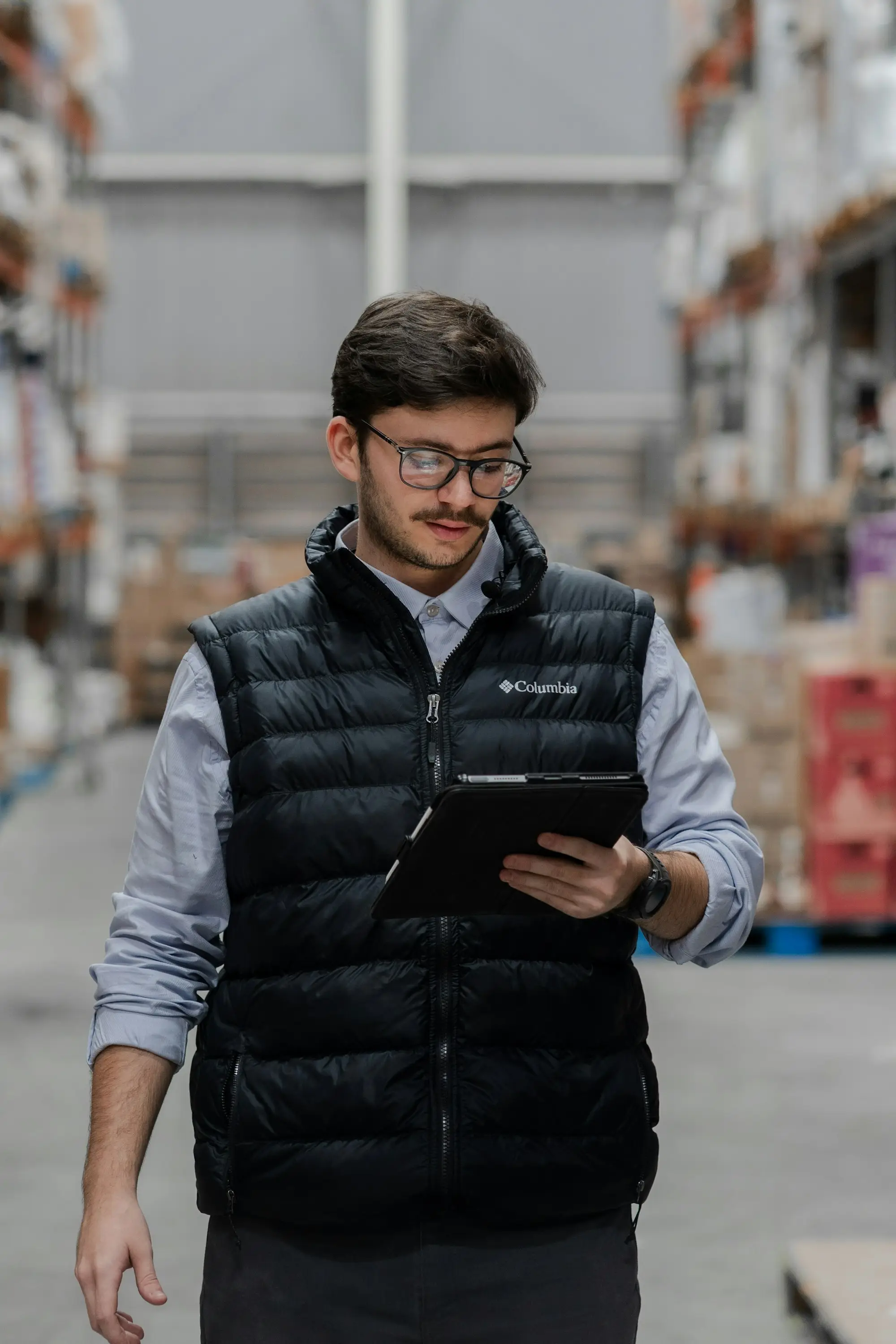 Warehouse manager in a puffer vest checking stock on a tablet between tall shelving aisles